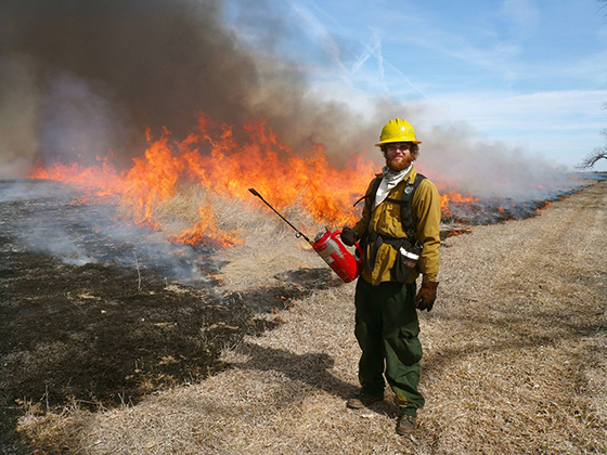 A person holding fire gear and dressed in yellow safety gear smiles and the camera as fire burns in a field behind them.