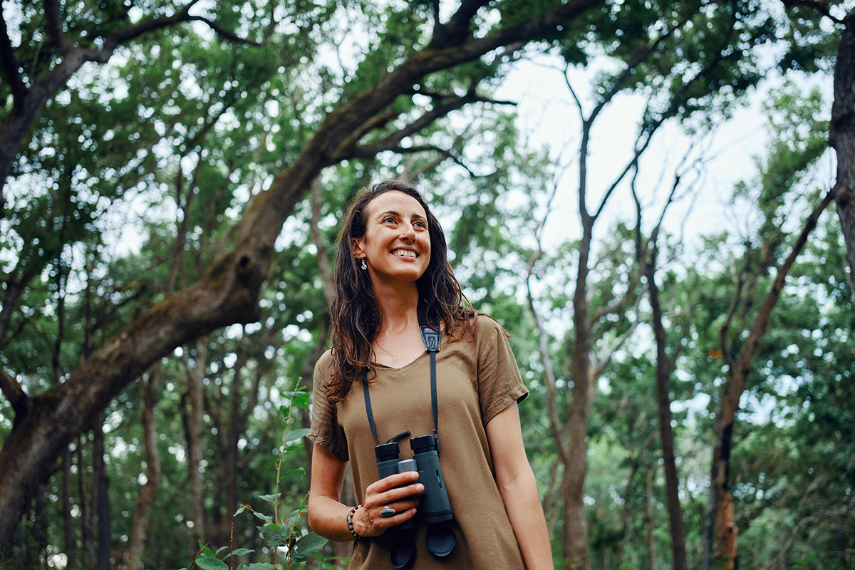 Bioaoustics researcher Maia Persche stands with binoculars in a Wisconsin forest. © Caleb Santiago Alvarado