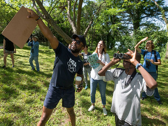 Several people in a field hold binoculars and point excitedly to the left.