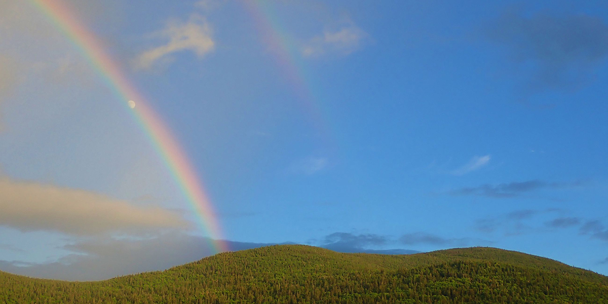 A bright rainbow arches over a bright blue sky and pale white moon before disappearing behind a lush green mountain top.