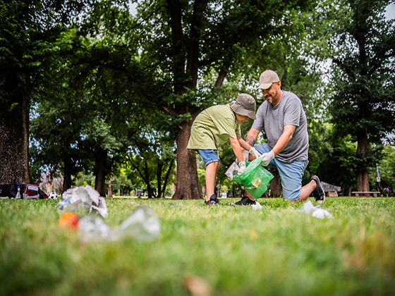 A father and son kneel in the grass as they pick up trash at a park.